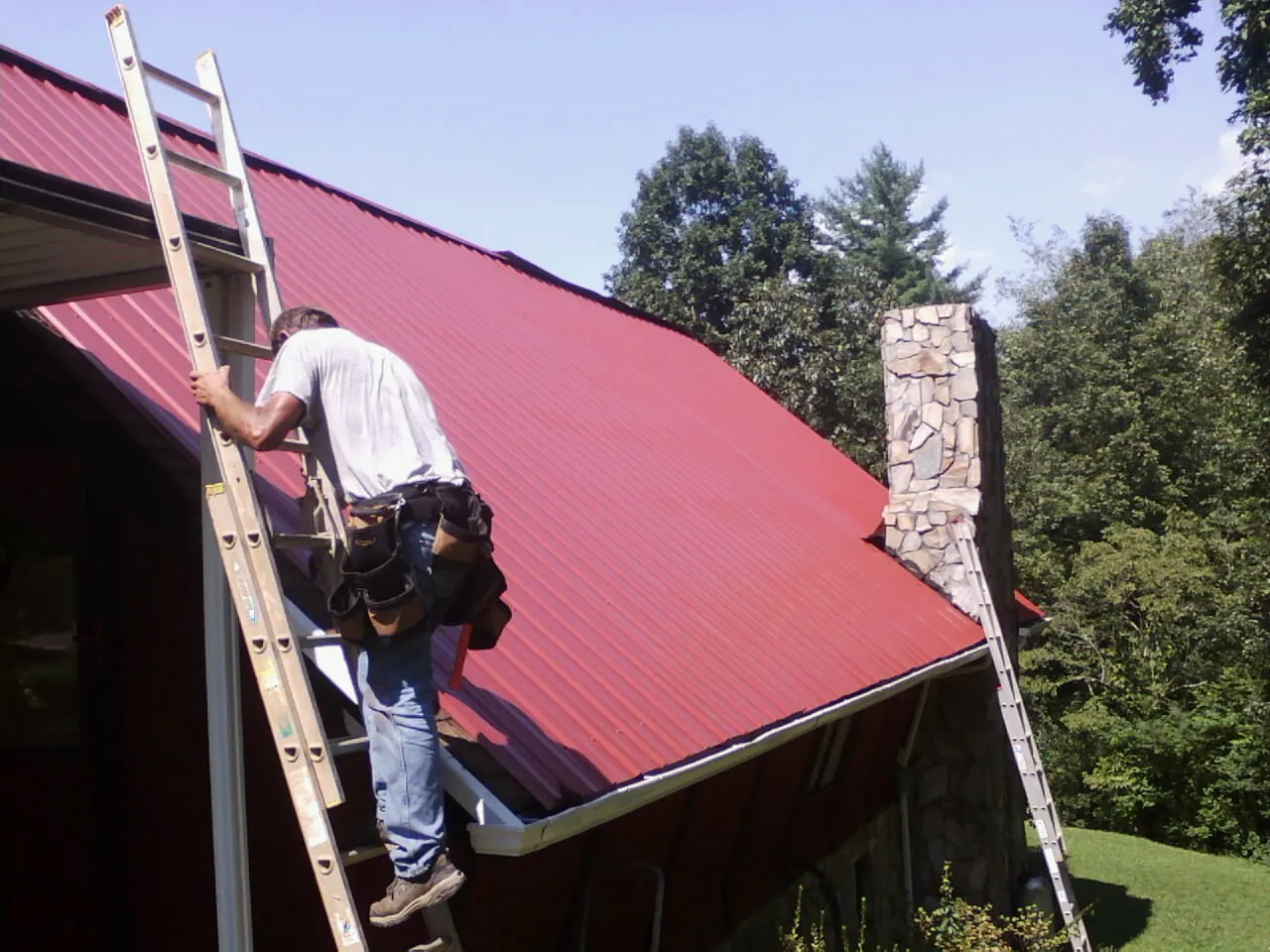 Roofer on a ladder beside a red metal roof and stone chimney — Soffit & Fascia Repair by Roofing Brothers in Copperhill